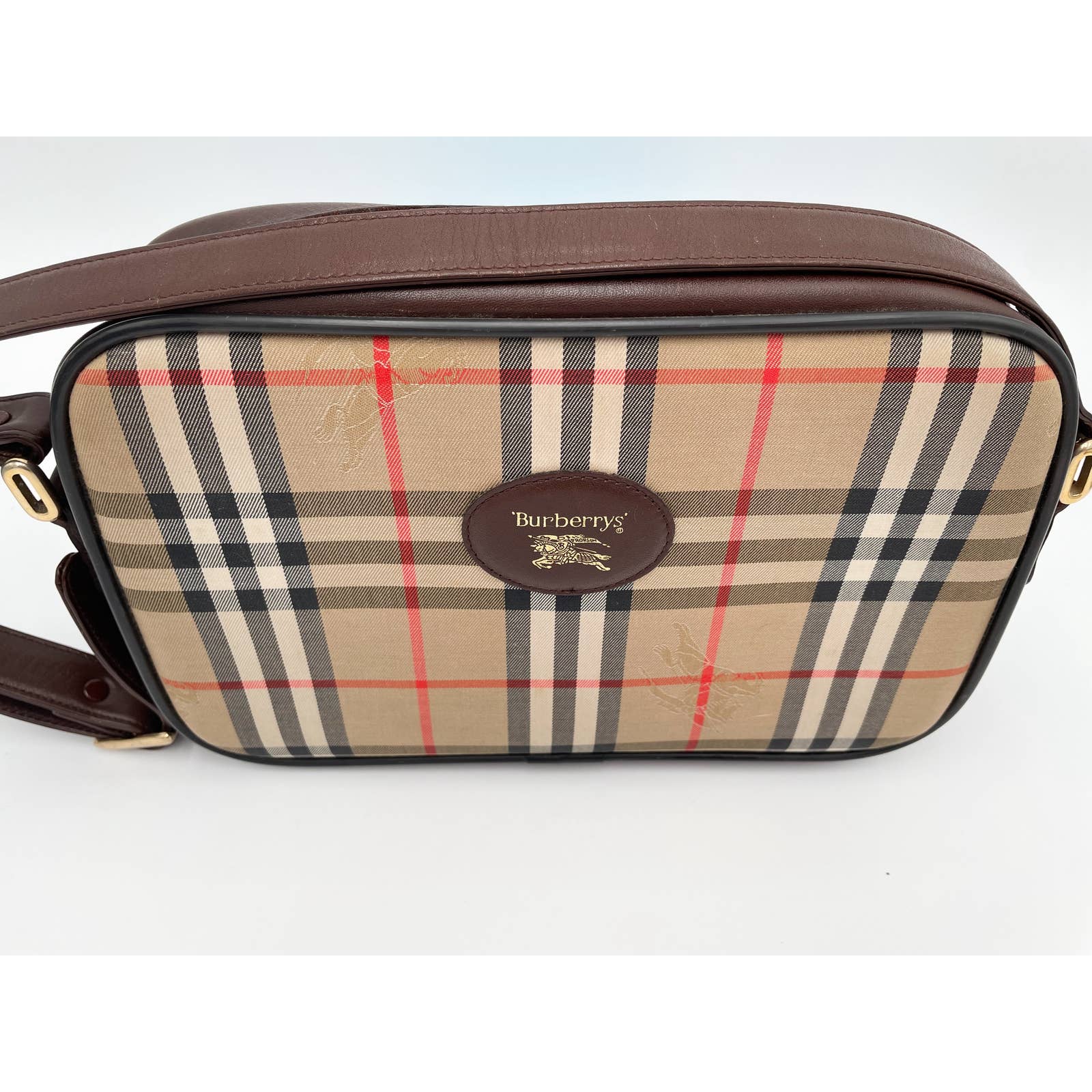 A rectangular vintage Burberry Crossbody Bag featuring the classic beige, black, white, and red pattern. The bag has a brown leather trim and adjustable strap, with an oval brown leather patch on the front displaying the "Burberry's" logo and equestrian knight emblem.