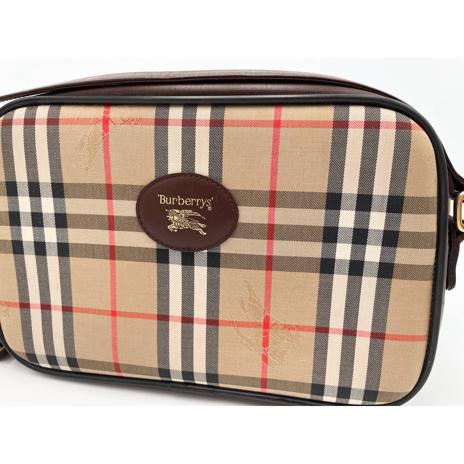 A rectangular vintage Burberry Crossbody Bag featuring the classic beige, black, white, and red plaid pattern. The center has a brown leather oval patch with gold-embossed text "Burberrys" and the equestrian knight logo. The bag has dark brown leather edges, a gold buckle, and an adjustable strap.