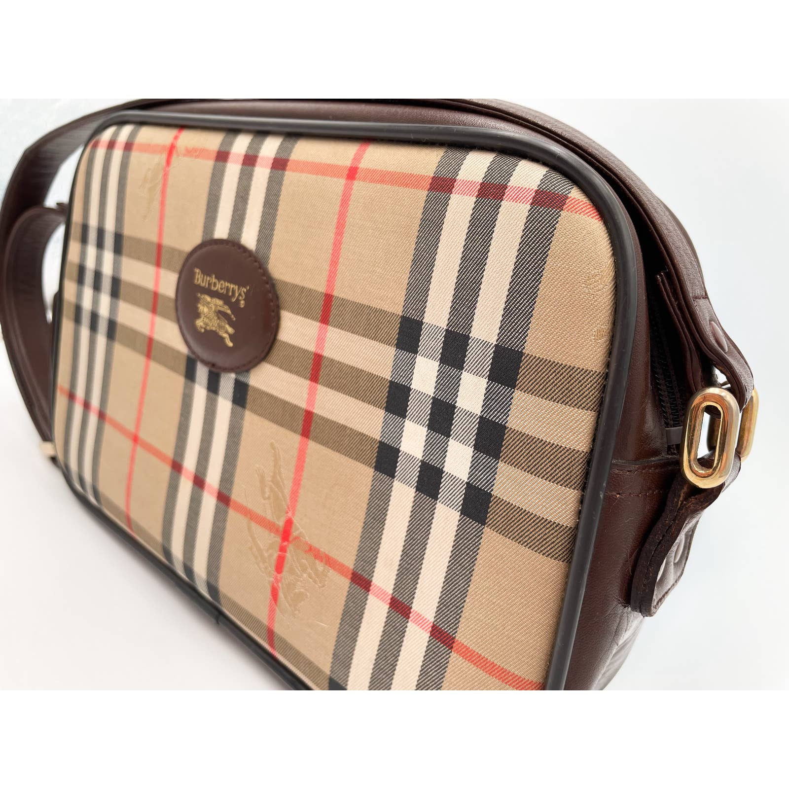 A Burberry Crossbody Bag featuring the brand's signature beige, black, red, and white plaid pattern. The bag has dark brown leather wear and an adjustable strap, with a close-up view of the Burberry logo embossed on a leather patch on the front.