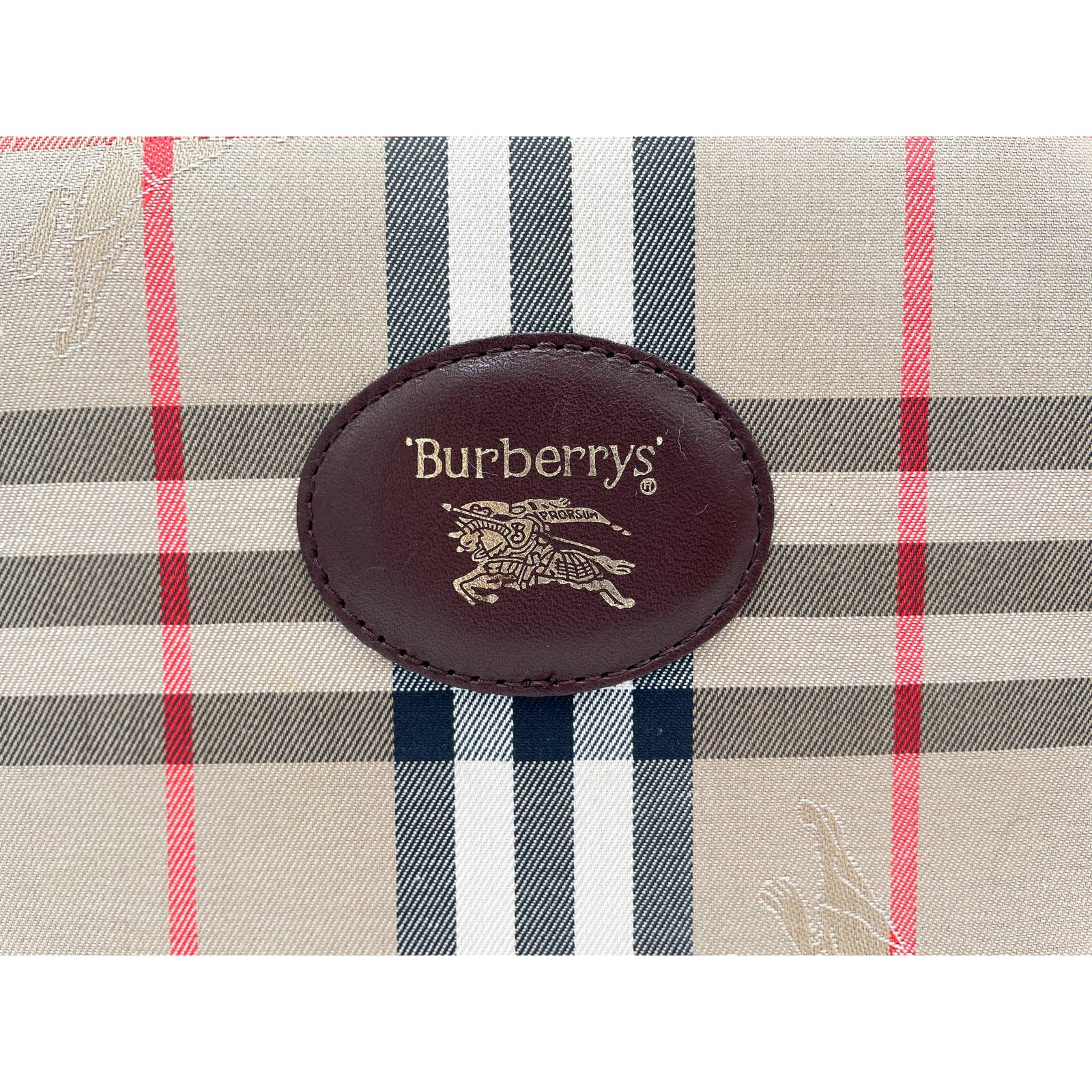 A close-up image of a beige fabric with a classic checkered pattern in red, black, and white, part of a Burberry Crossbody Bag. At the center, there's a brown leather oval patch with the text "Burberrys" and a knight logo embossed in gold.