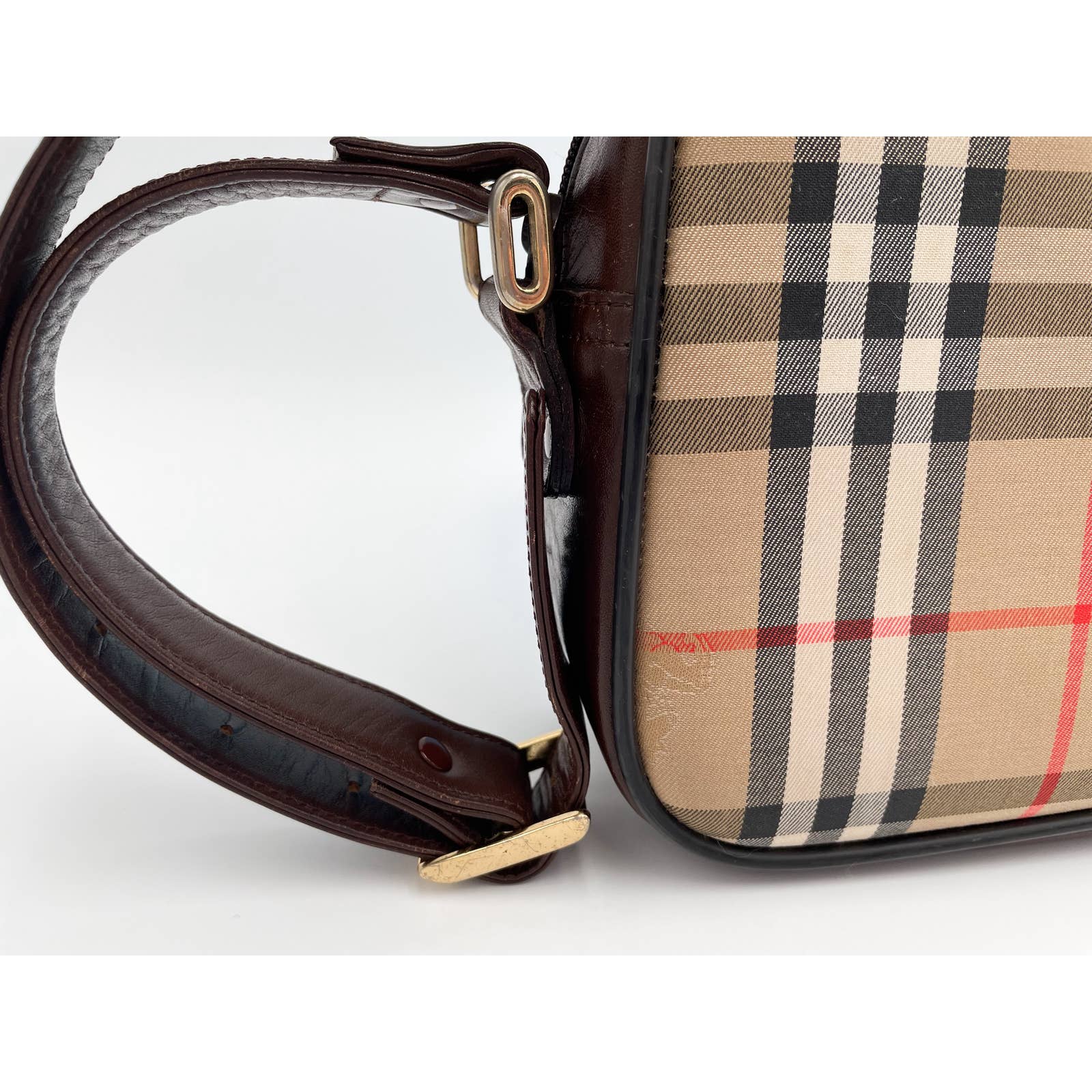 A close-up of a Burberry Crossbody Bag with a checkered pattern featuring brown, black, white, and red stripes. The bag has a brown leather strap with a gold buckle and an adjustable strap. A small golden clasp adds a touch of elegance. The background is white.