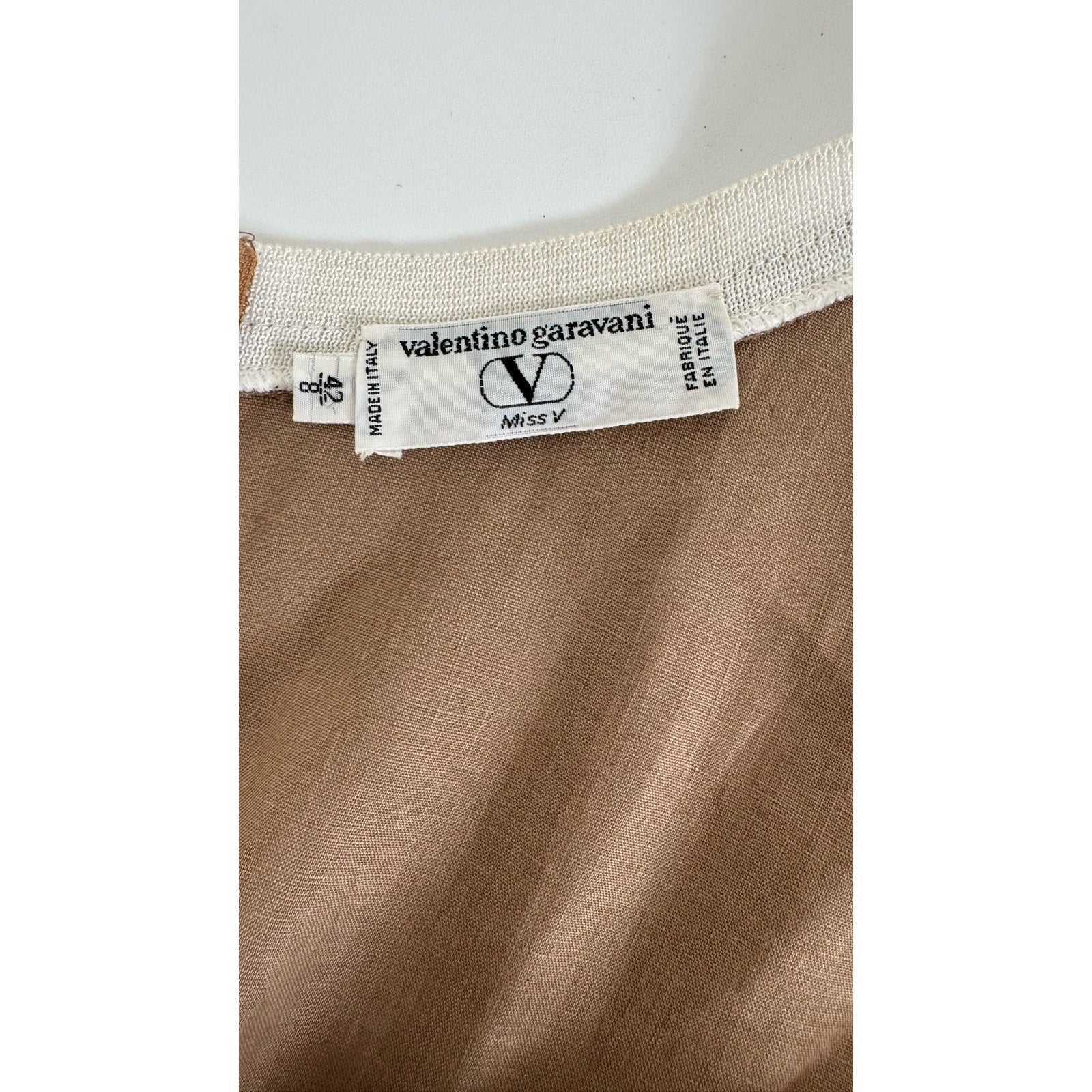 Close-up image of a clothing label with "Valentino Miss V" written on it, along with "Made in Italy" and "Fabrique en Italie." The label is attached to a light brown fabric with white stitching along the seam, hinting at a vintage Valentino Miss V Linen Tank Top crafted from 100% linen.