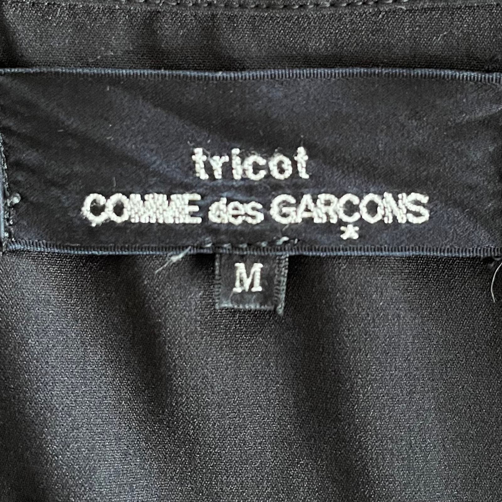 Close-up of a clothing label showing the brand "Comme des Garçons" in white text on a black background. Below the brand name, there is a small tag with the letter "M" indicating the size medium. The polyester fabric around the label is also black. The product shown is a Vintage 1992 Comme des Garcons Asymmetric Top.