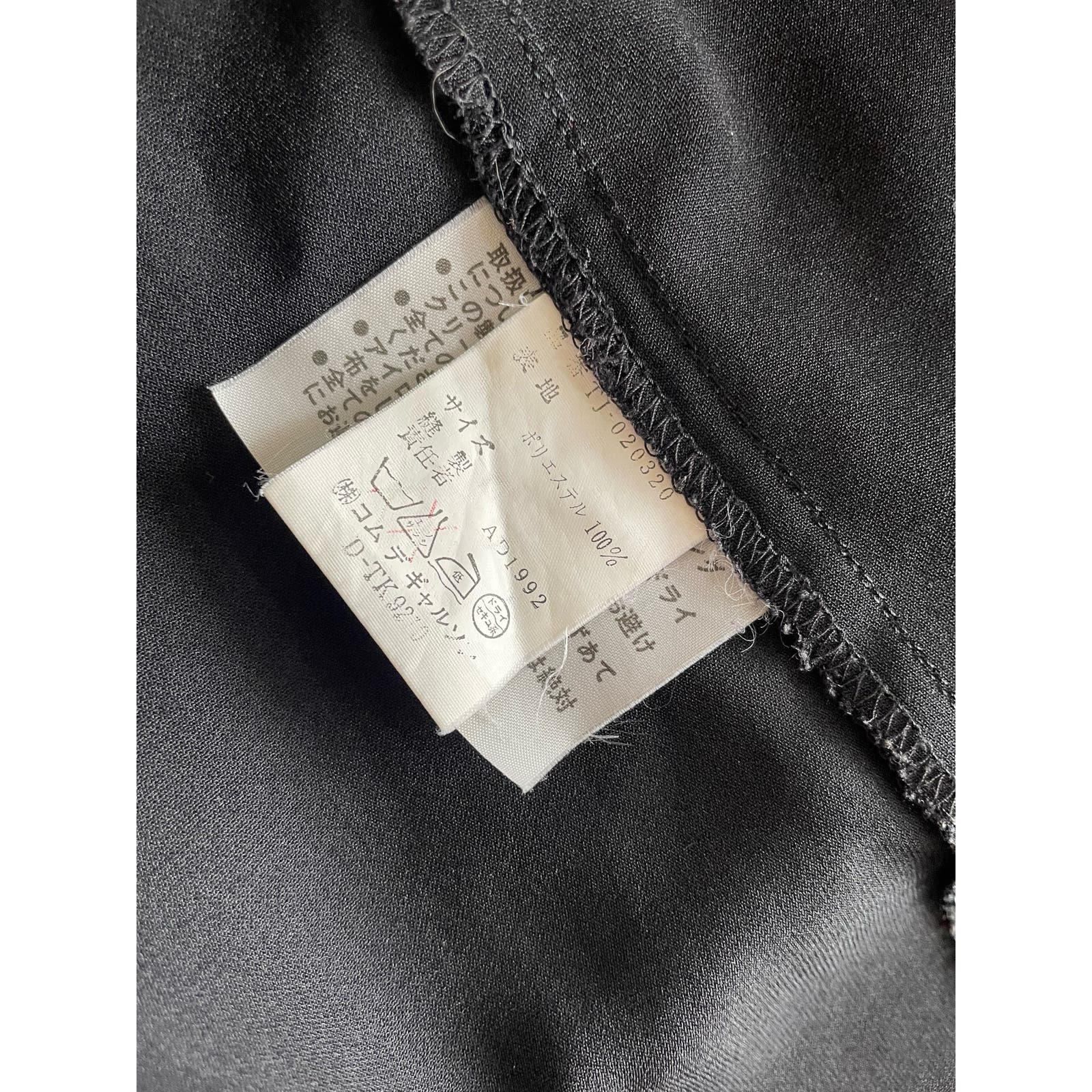 Close-up of a garment care label with text in Japanese, including care instructions and fabric composition details. The label, from the renowned brand Comme des Garçons, is attached to a piece of black polyester fabric, showing clear stitching and seams around it. This particular piece is the Vintage 1992 Comme des Garçons Asymmetric Top.