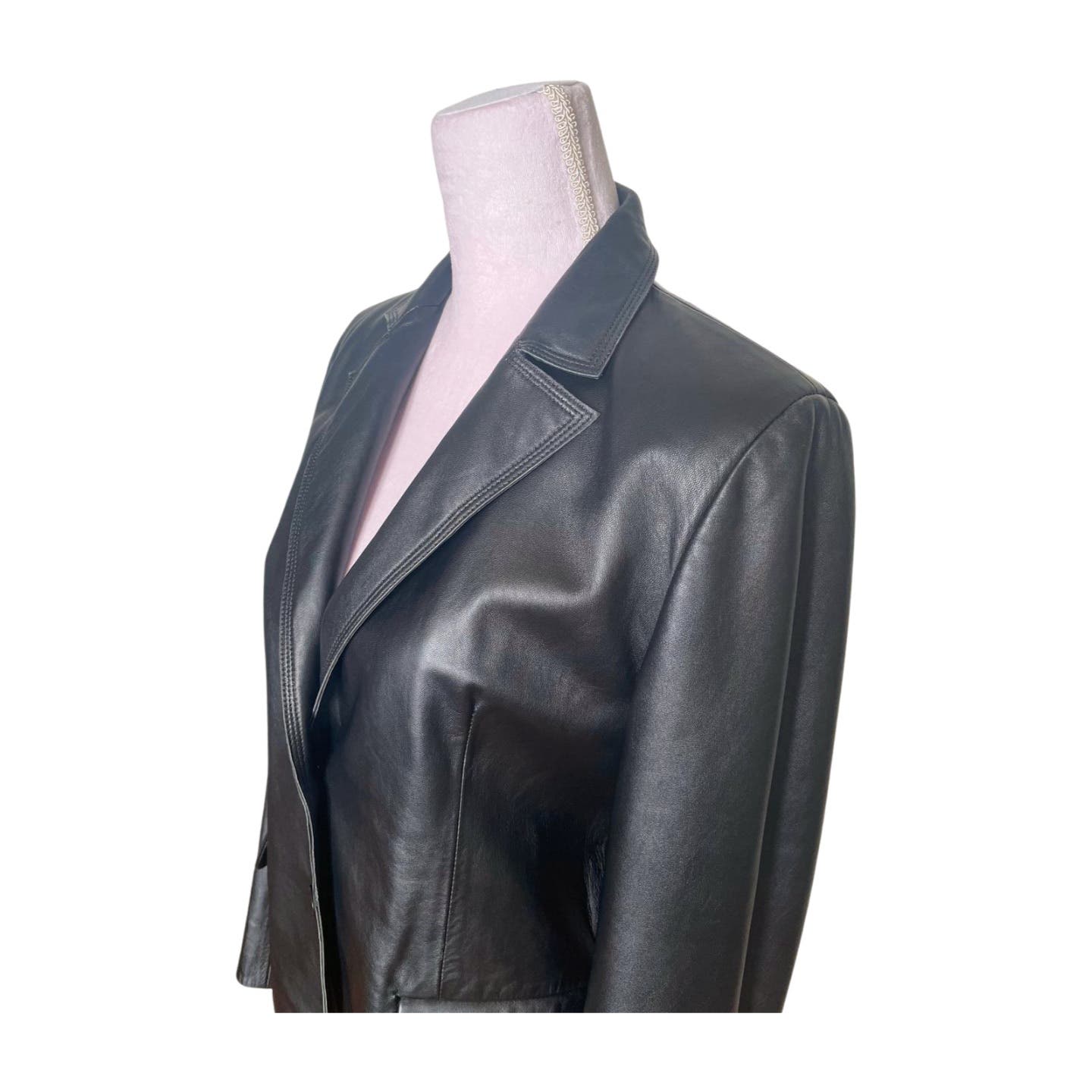 A Vintage Balmain Black Leather Blazer displayed on a pink dress form. The blazer features a notched collar and a sleek, fitted design with a smooth, polyester lining. The image focuses on the upper portion of the blazer, highlighting its smooth texture and tailored seams. The background is plain white.