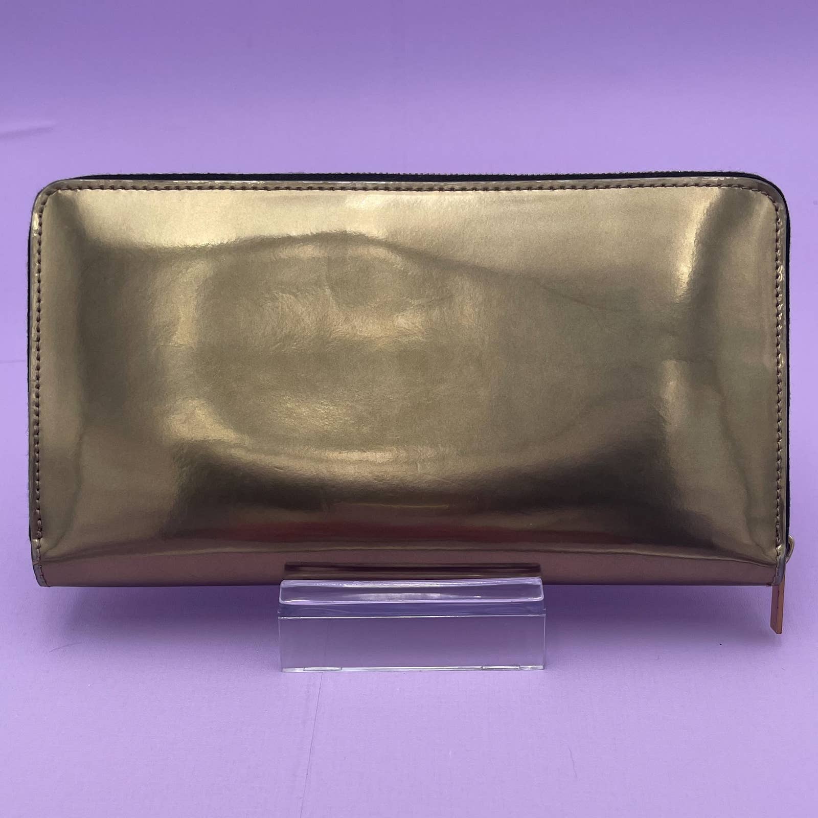 The image shows a shiny, metallic gold Celine Alphabet Wallet Gold with a zipper closure. Made of patent leather, the wallet is standing upright on a clear stand against a lavender background.