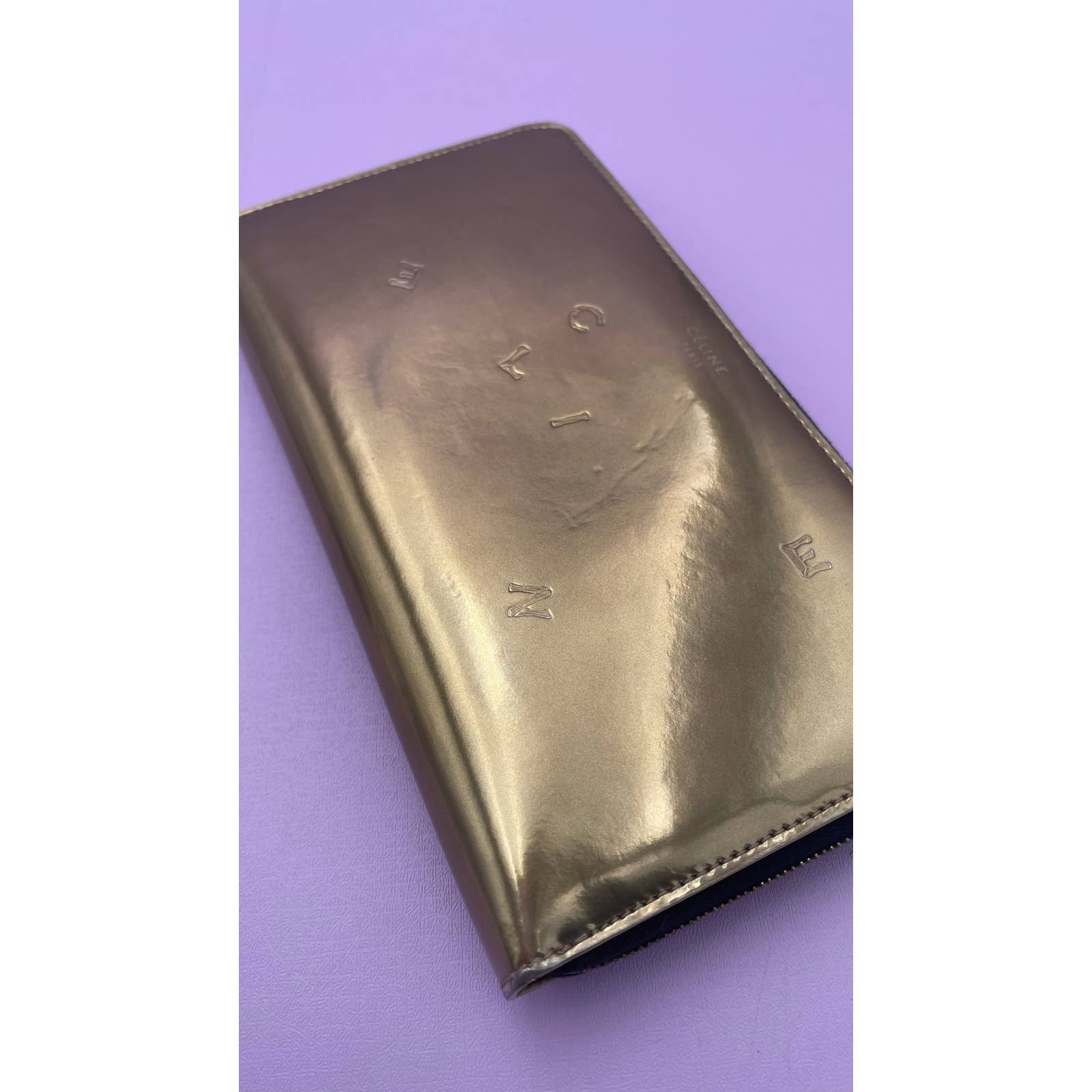 A shiny, gold-colored Celine Alphabet Wallet Gold in patent leather with a zipper closure rests on a purple background. The wallet features embossed letters and symbols on its surface.