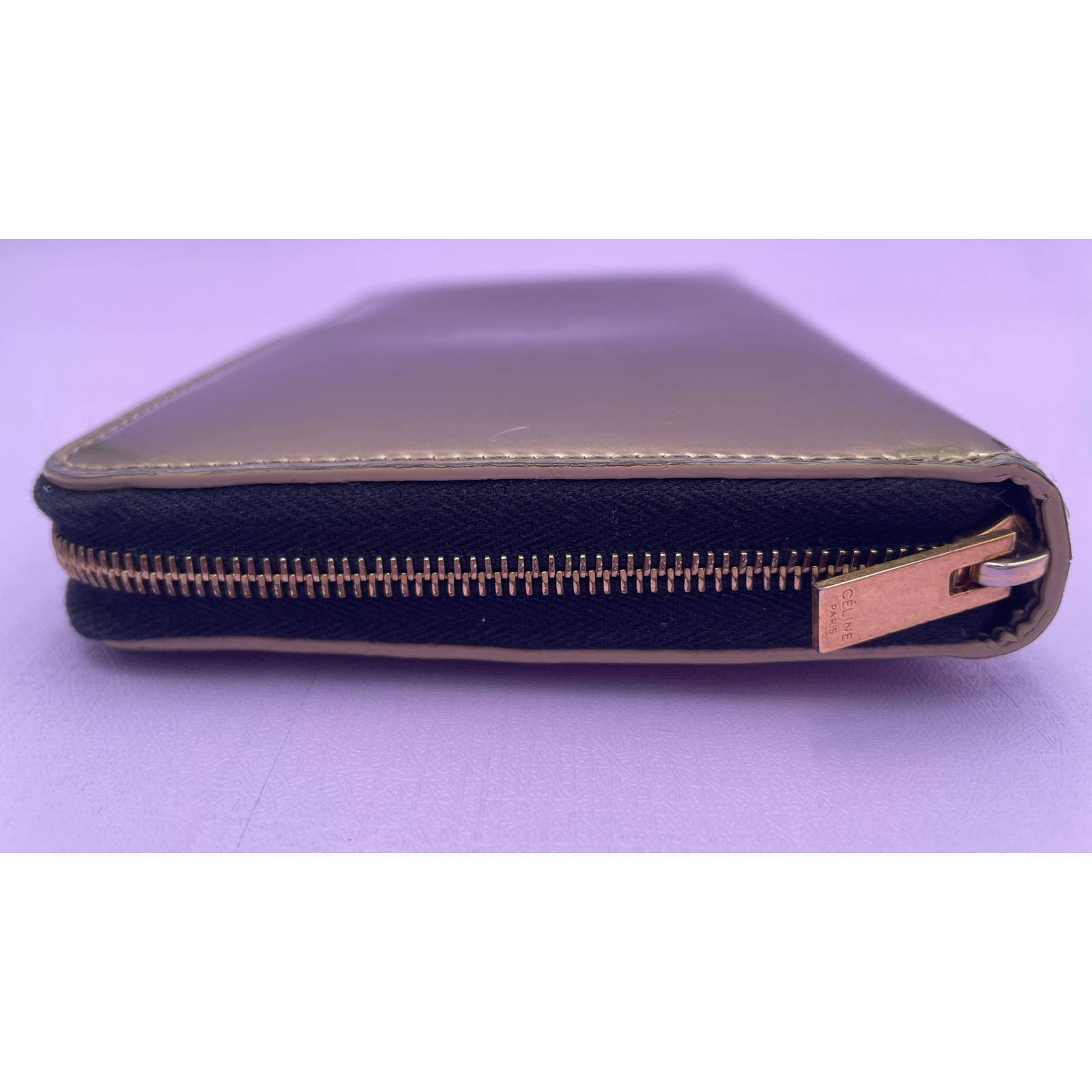 A close-up view of the Celine Alphabet Wallet Gold, made from patent leather, positioned horizontally with the zipper closure slightly open. The wallet is dark with a golden zipper and laid on a light purple surface.