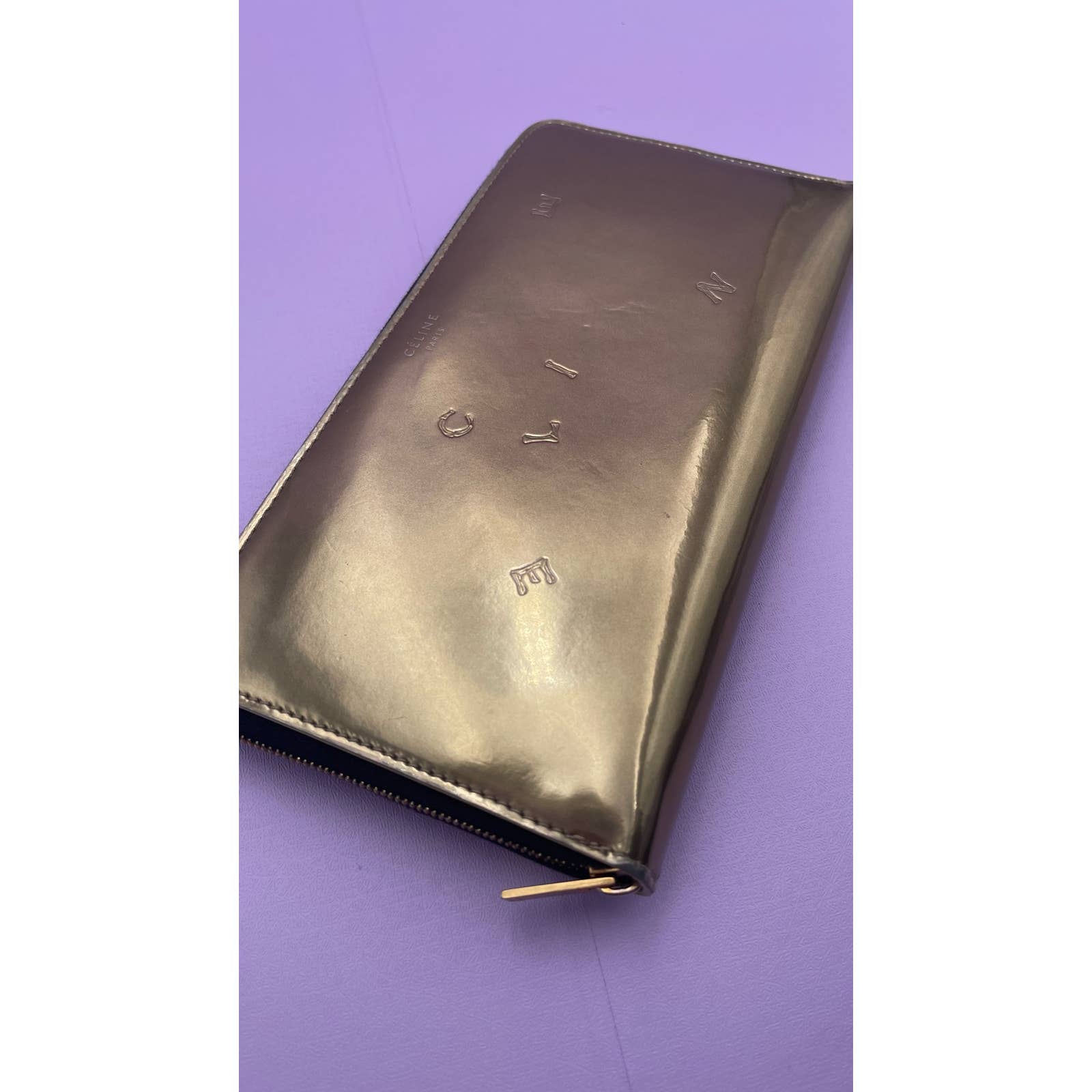 A sleek, metallic Celine Alphabet Wallet Gold lies flat on a pastel purple surface. The patent leather wallet reflects light, showing a lustrous finish. Stamped letters are subtly visible on its surface, but the full text is not entirely clear.

